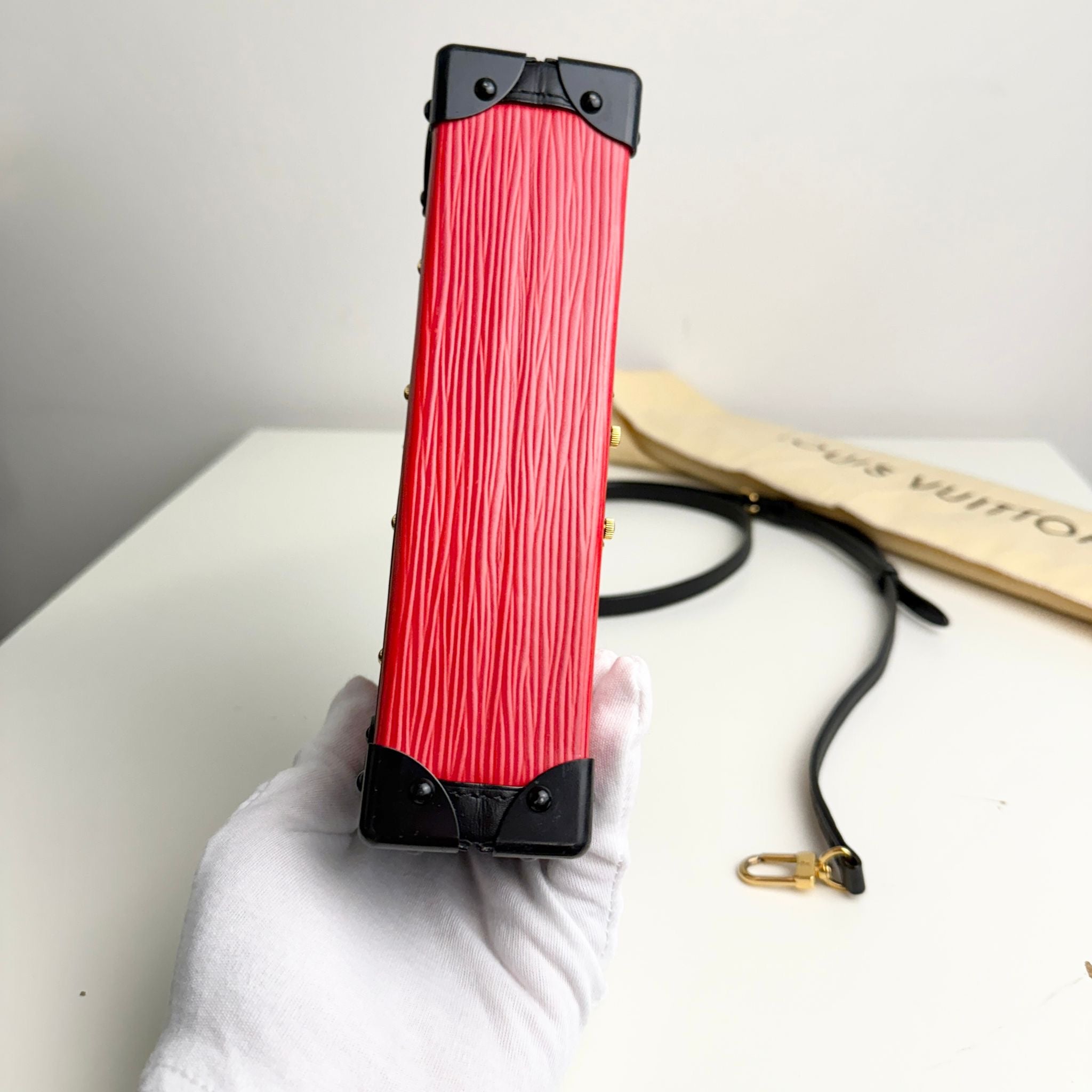 Red and black rectangular device held by a gloved hand on a white surface with a Louis Vuitton box in the background.