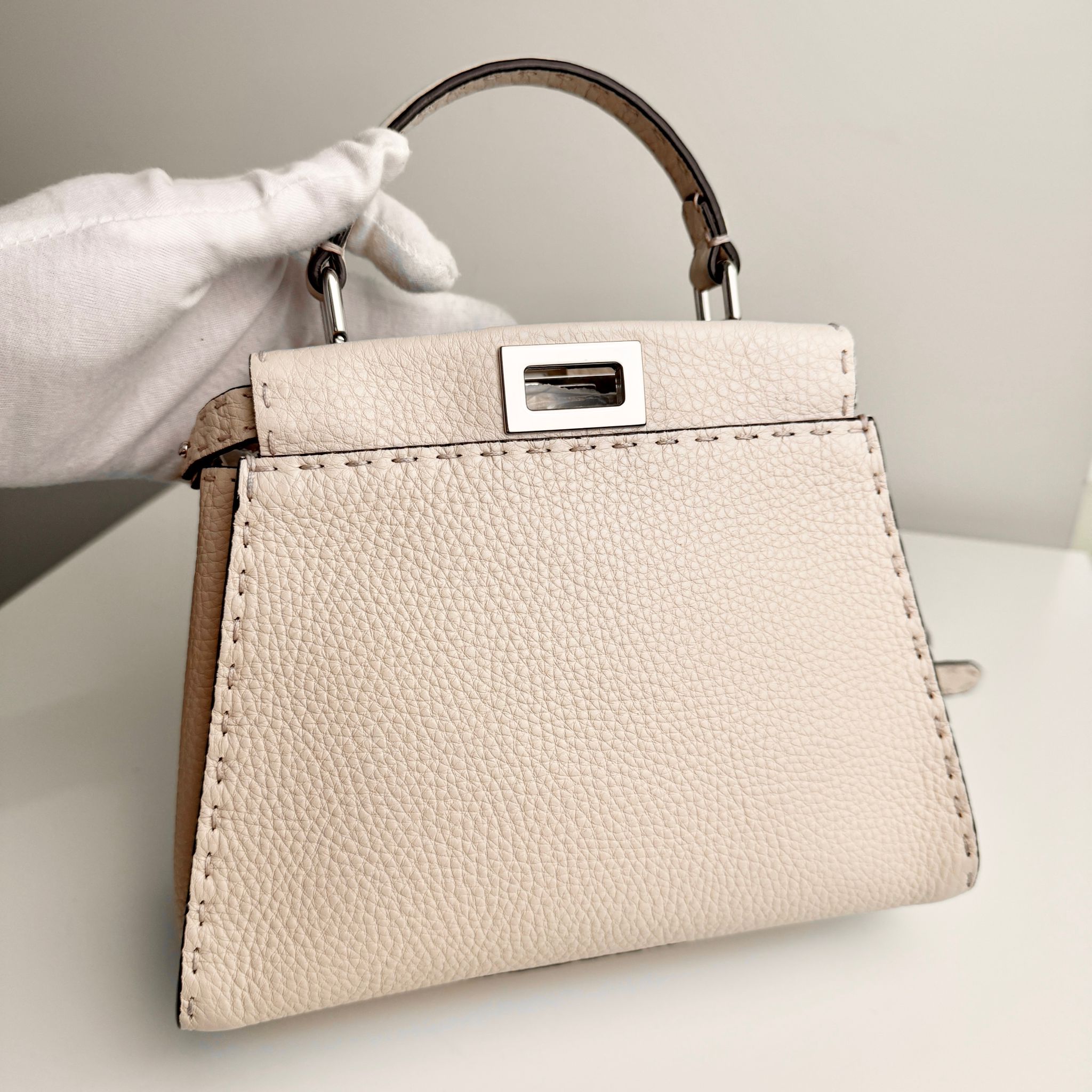 Beige handbag with a silver clasp held by a gloved hand on a light background