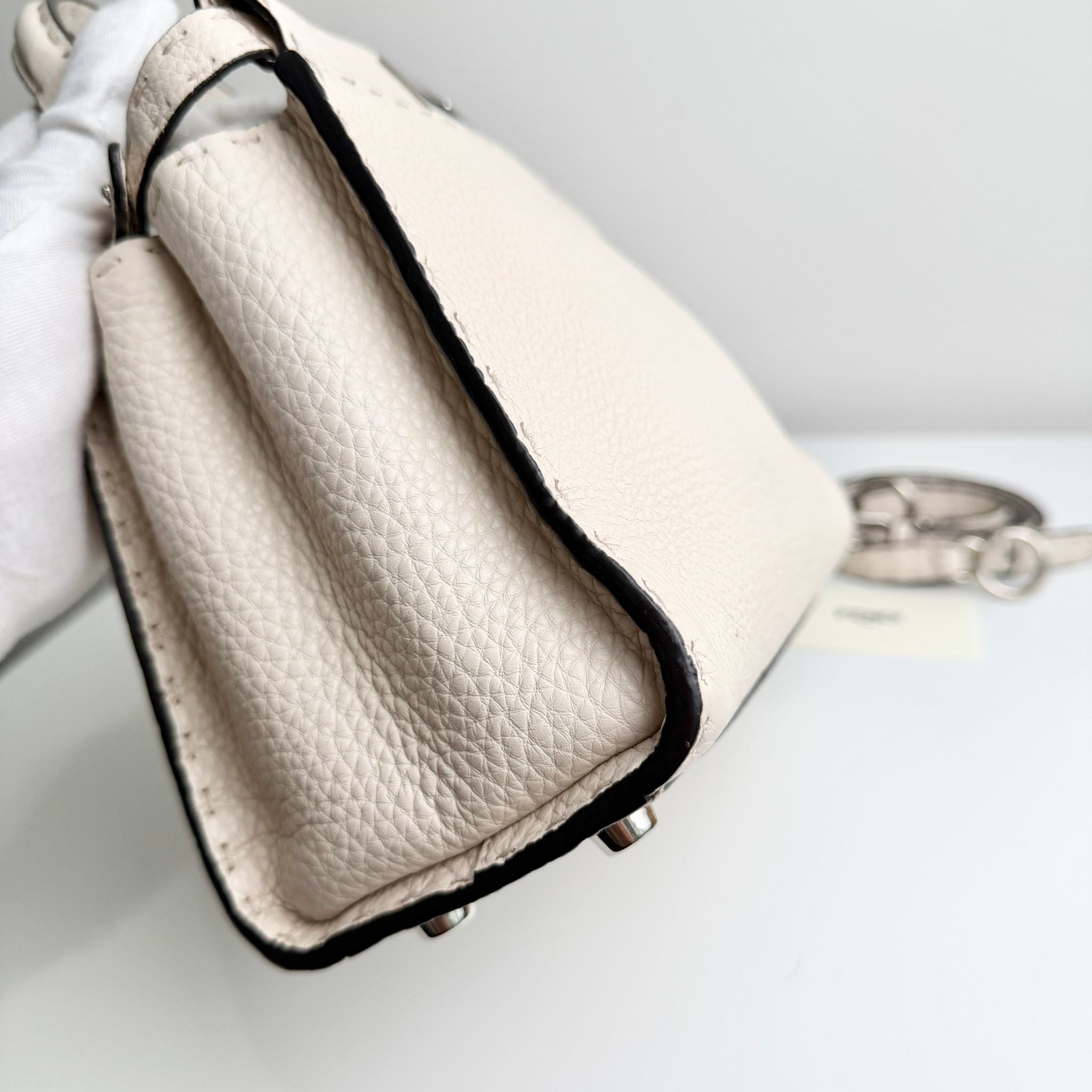 Close-up of a beige handbag with black trim held by a gloved hand on a light gray background.