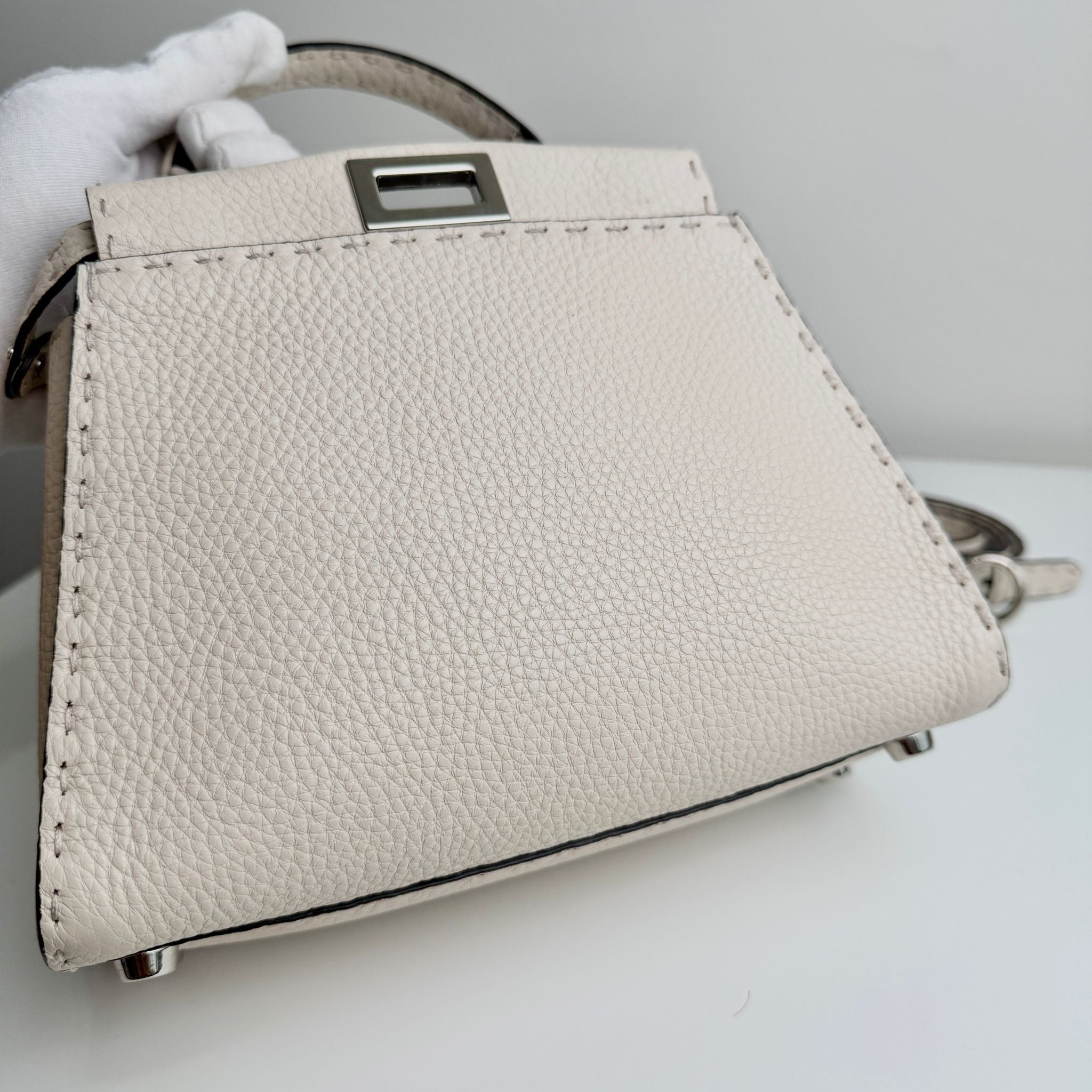 Beige handbag with a visible brand logo held by a gloved hand on a light gray background