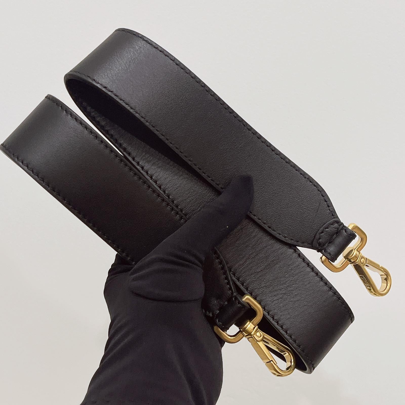 Black leather bag strap with gold hardware held by a gloved hand on a light gray background