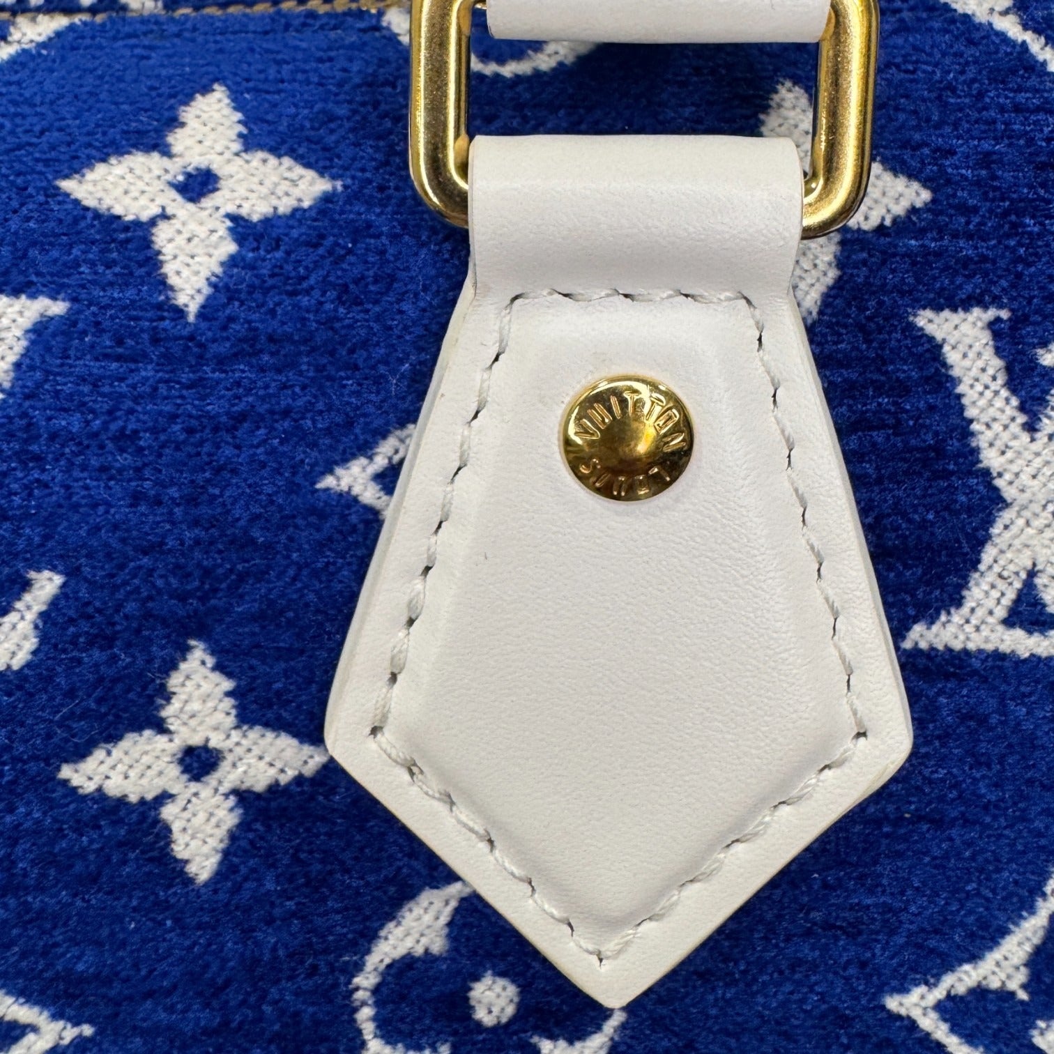 Close-up of a white leather strap with gold hardware on a blue fabric background with brand logo.