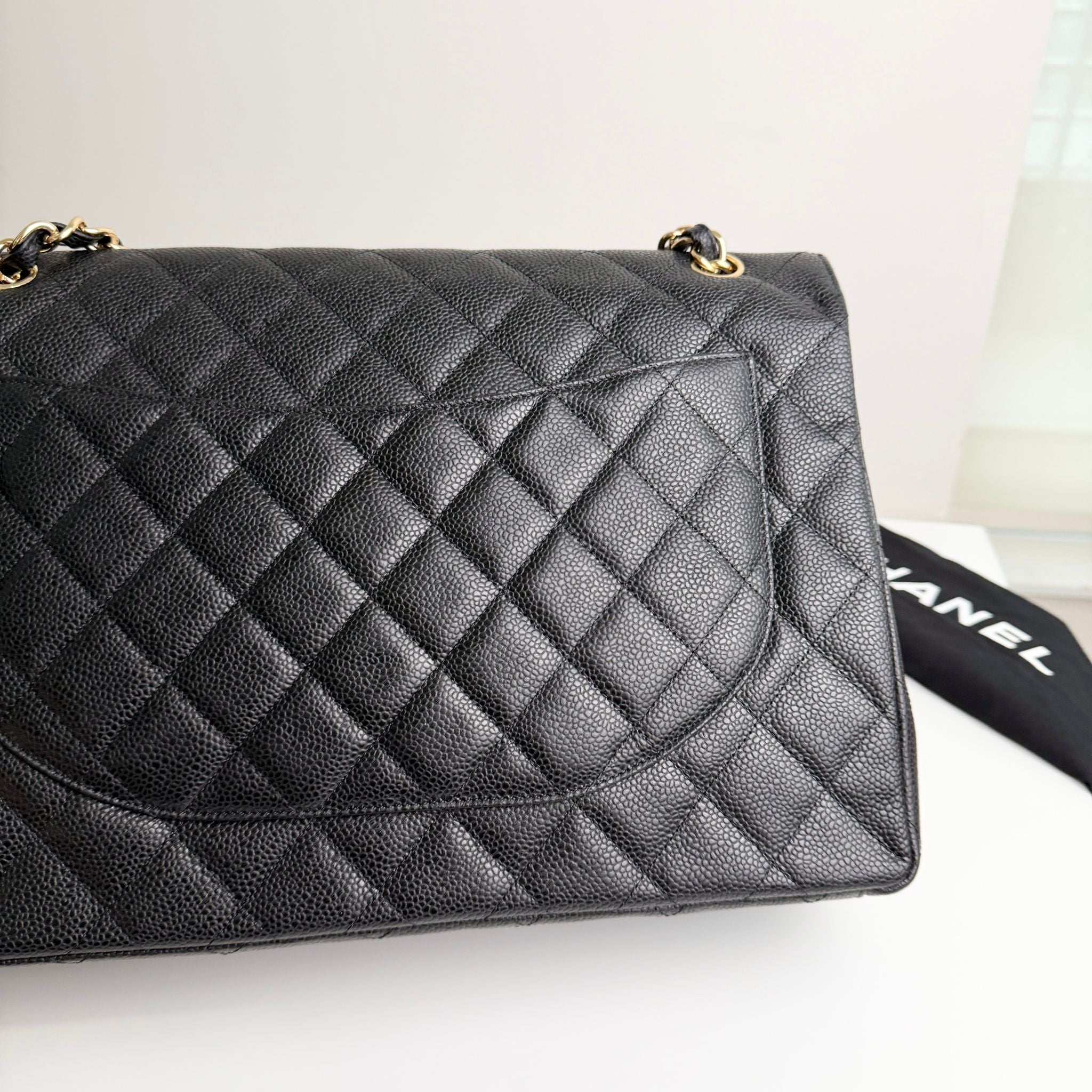 Black quilted handbag with gold hardware on a white surface, with a black Chanel dust bag partially visible.