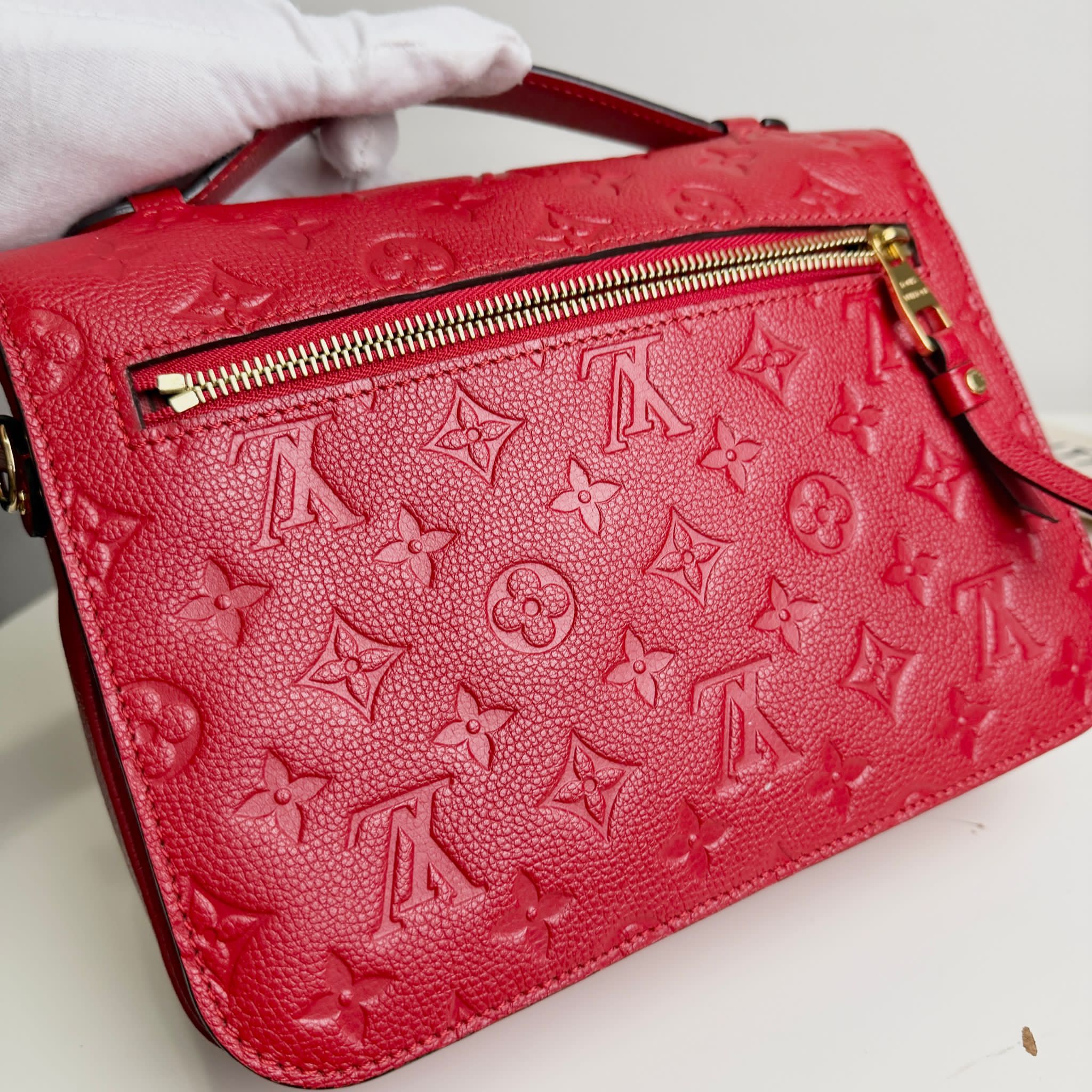 Red handbag with a prominent logo pattern held by a gloved hand on a white background
