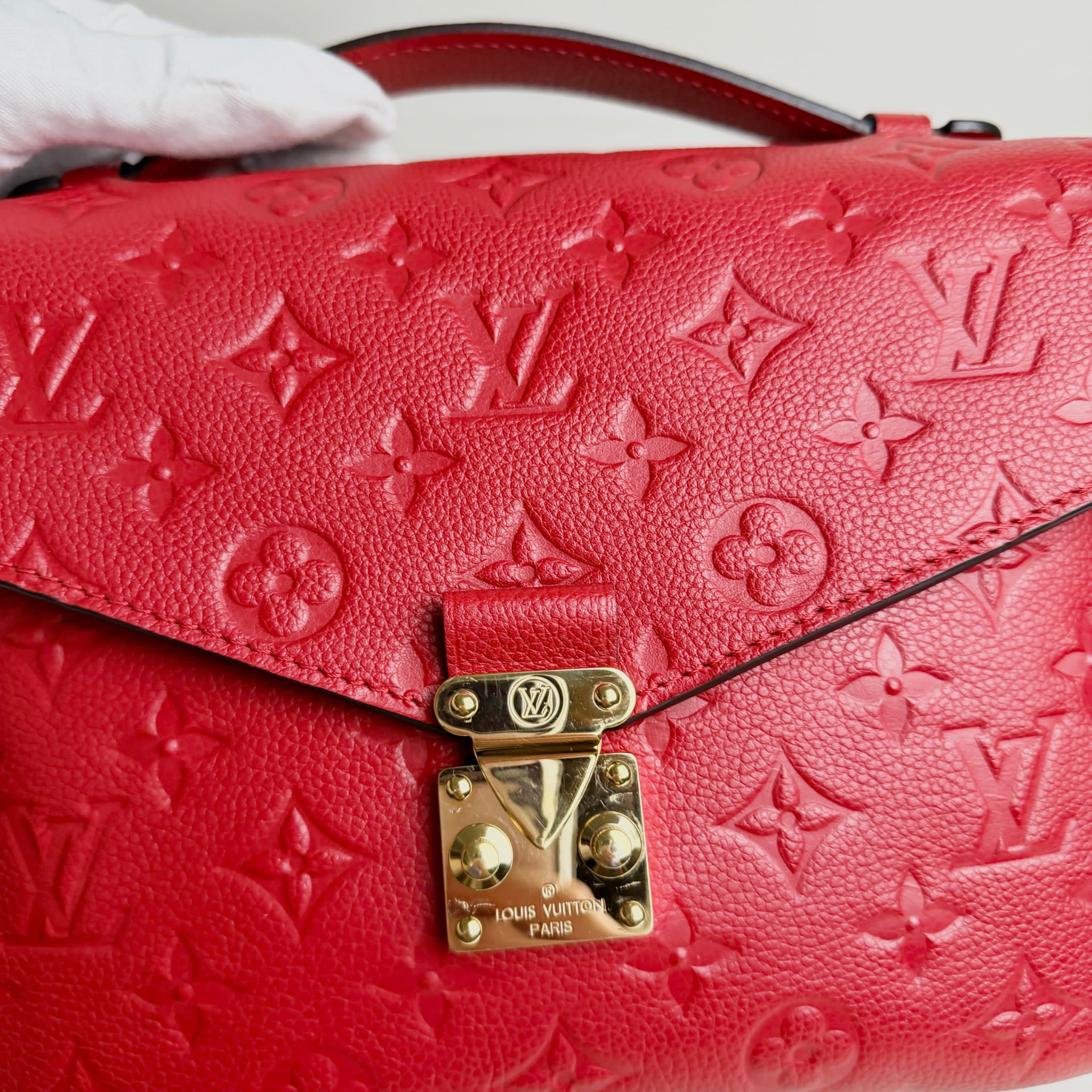 Red handbag with gold lock and Louis Vuitton logo, held by a gloved hand.