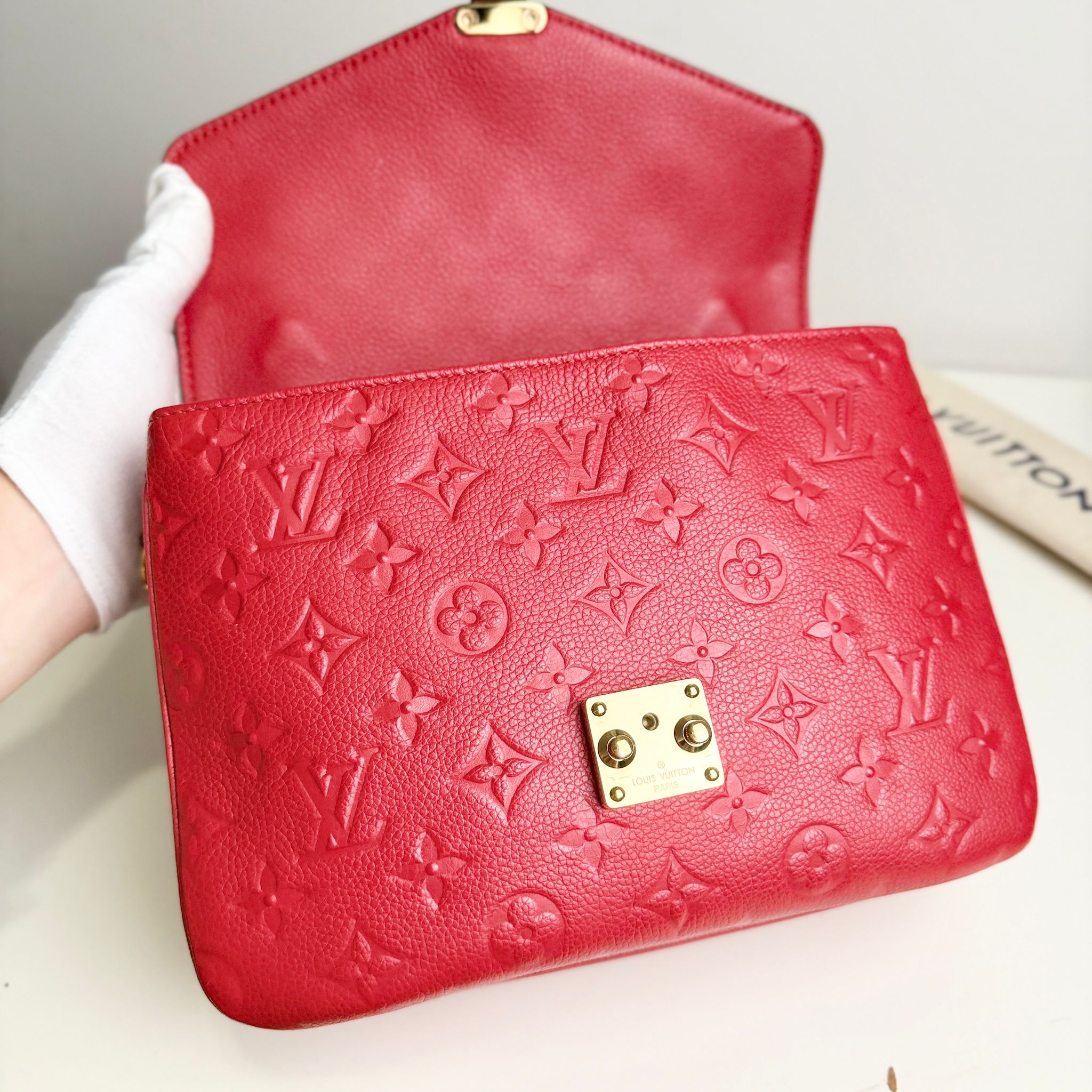Red wallet with gold accents and monogram pattern held by a gloved hand.