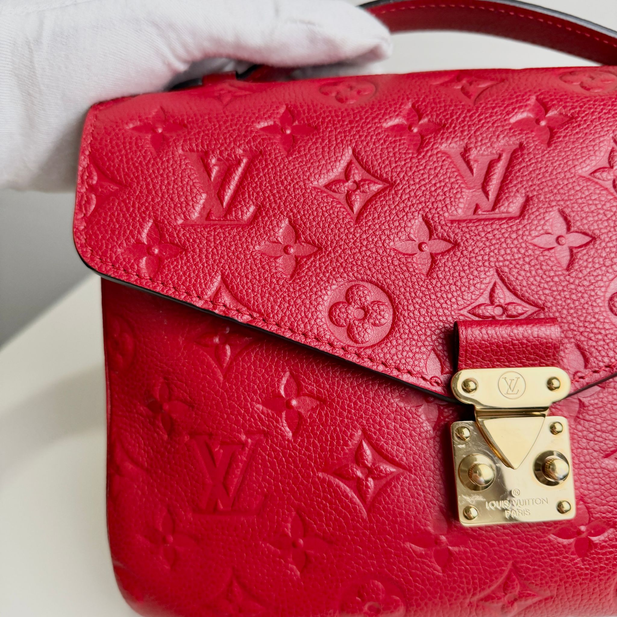 Red handbag with gold lock and monogram pattern, held by a gloved hand.